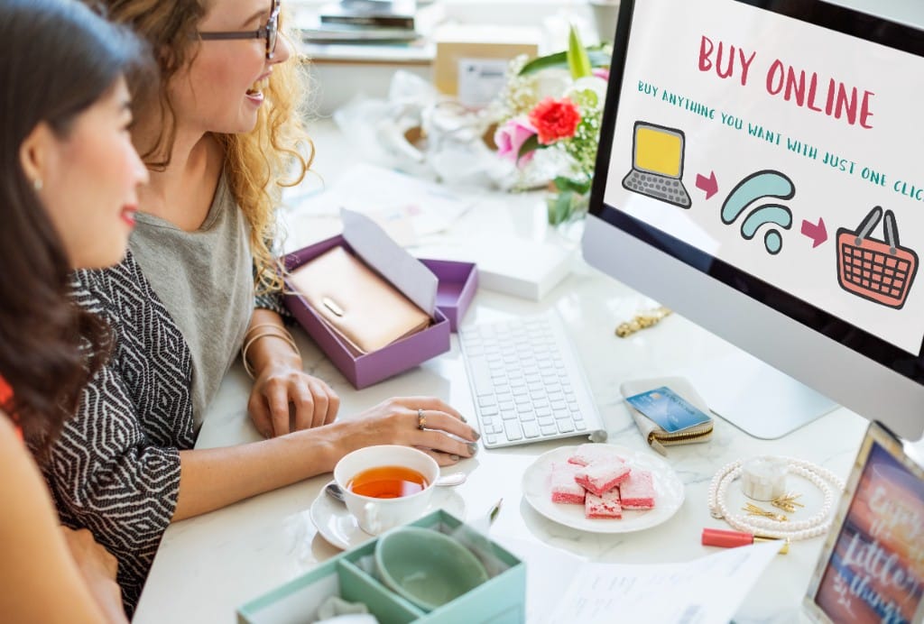 Two women smiling at a computer screen showing a Buy Online ecommerce graphic with a credit card on the desk