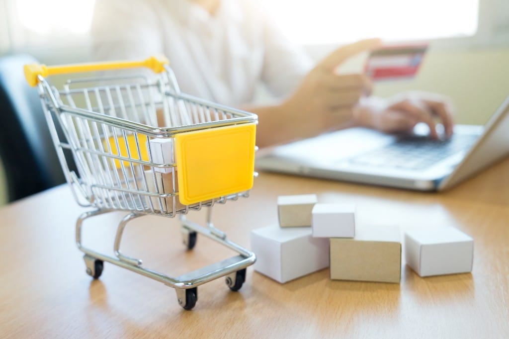 Miniature shopping cart and delivery boxes on a desk with a person paying by credit card on a laptop in the background