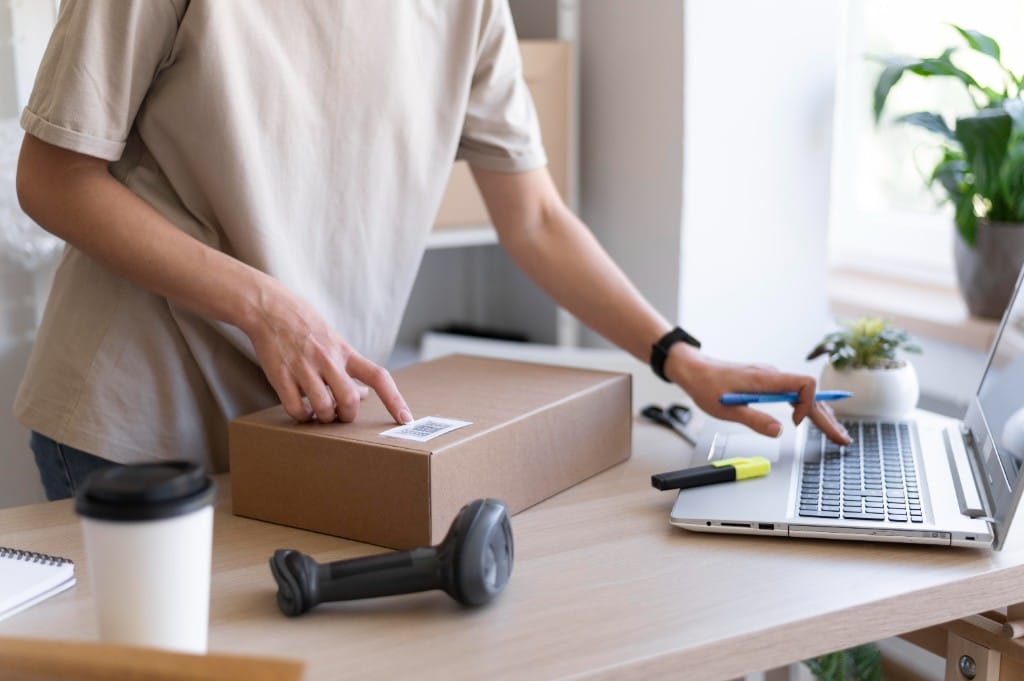 Person managing ecommerce orders on a laptop while preparing a shipping box with a barcode scanner on a desk