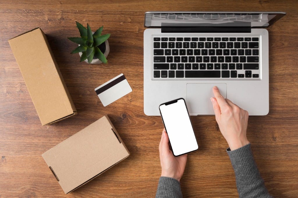 Top-down view of a person using a laptop and smartphone for online shopping beside cardboard shipping boxes and a credit card