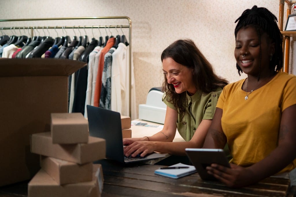 Two women collaborating on ecommerce order fulfillment with a laptop and tablet in a clothing warehouse