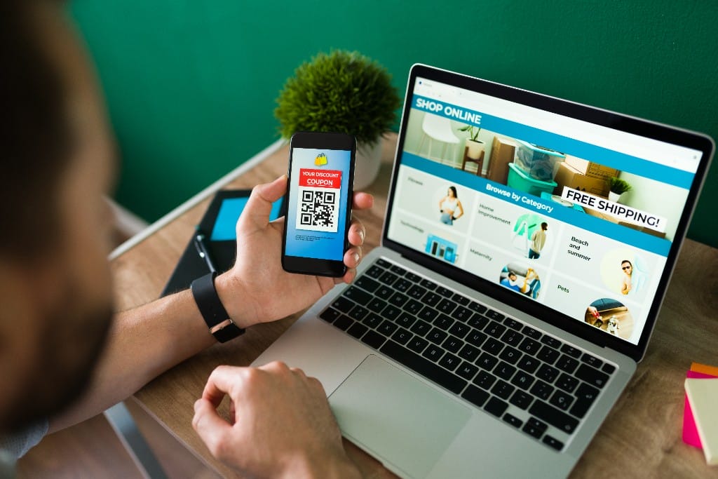 Person browsing a modern ecommerce website on a laptop while holding a smartphone with a discount coupon QR code
